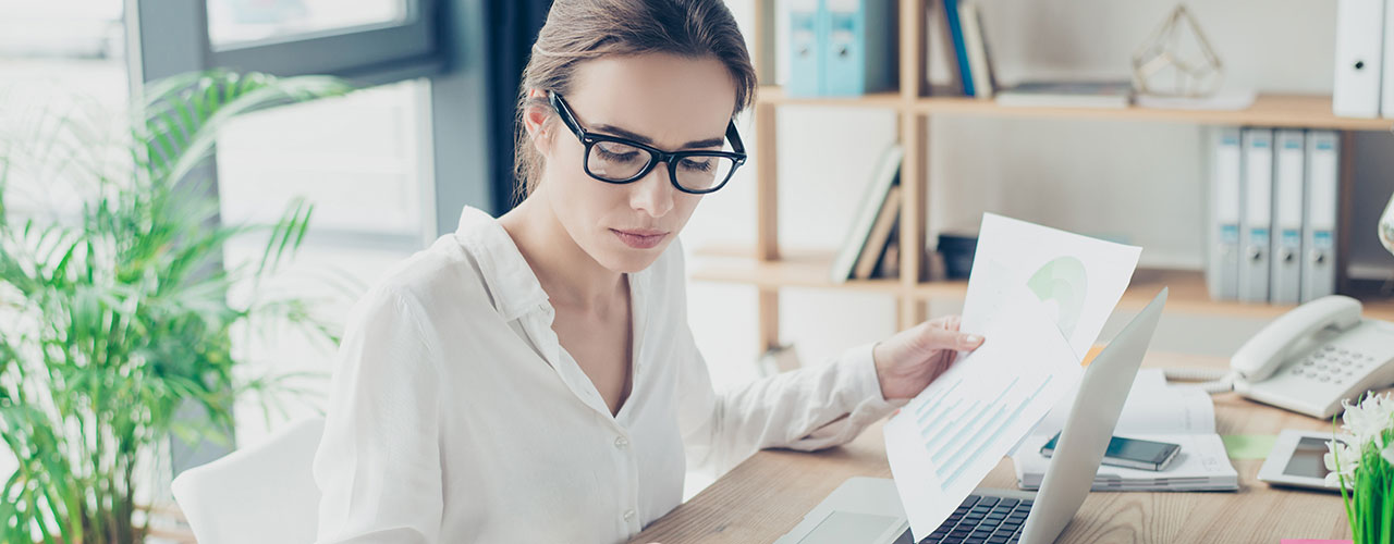 Geschäftsfrau in formeller Kleidung mit Brille sitzt am Arbeitsplatz in einem hellen, modernen Büro und macht einen Bericht mittels Taschenrechner vor einem Laptop
