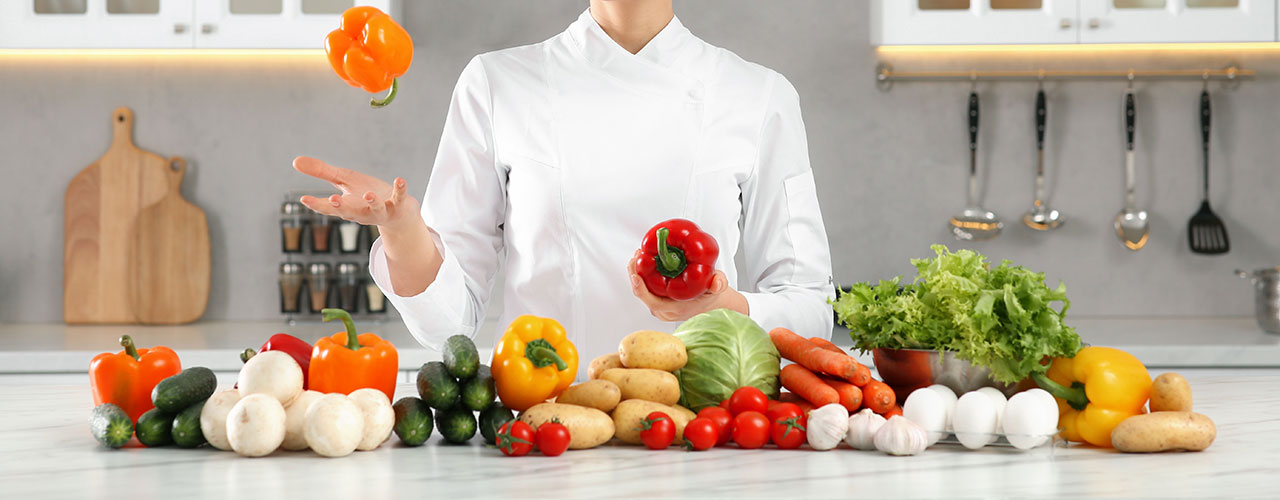 Glückliche Köchin wirft Paprika in die Luft und am Tisch in der Küche liegt frisches Gemüse