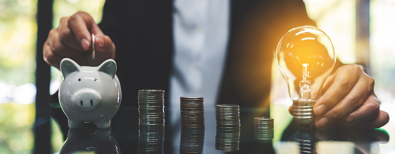 Eine Geschäftsfrau wirft Münzen ins Sparschwein, während sie eine Glühbirne in der Hand hält und Münzen auf dem Tisch stapelt